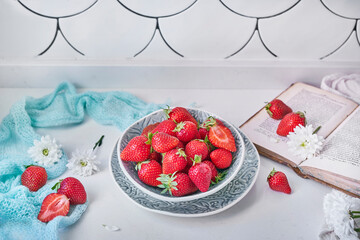 Bowl of strawberries near flowers and book on cabinet