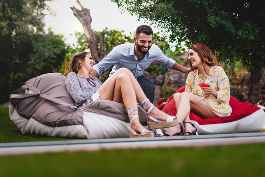 One Young Man And Two Women Talking And Laughing Carefree Sitting On Bean Bags At Luxury Backyard Party Drinking Fancy Cocktails