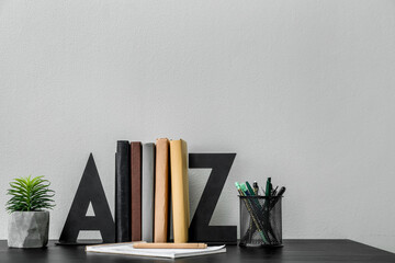 Stylish holder with books, pen cup and flowerpot on table near light wall
