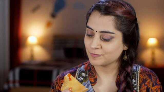 A young Indian woman in her twenties enjoying Samosa with tomato ketchup / Chutney - delicious food  deep-fried. A pretty Indian lady bingeing on unhealthy food - junk food  poor lifestyle  cheat d...