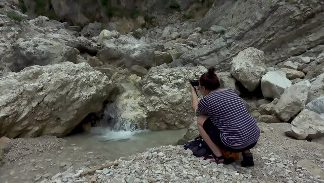 Hiker woman photographer in a rocky valley with spring stream water in the mountain river. Photographer hiker taking pictures of nature.