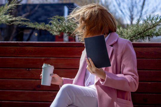 A young beautiful girl in sunglasses drinks coffee from an environmentally friendly thermos and holds a notebook in her hands, sitting on a bench outside in windy weather
