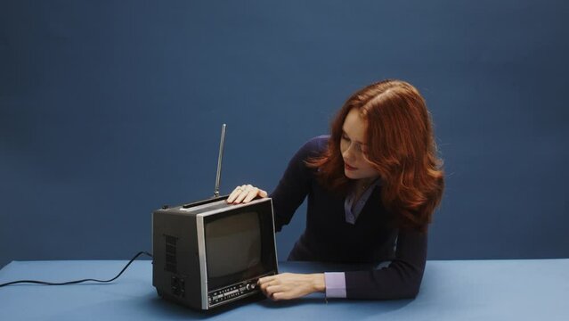 A young ginger white woman turning an old retro television on, getting popcorn and enjoying tv show, wearing a purple dress in front of a blue background sitting at a blue desk