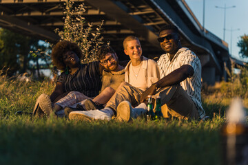 Fototapeta na wymiar Happy diverse group of young friends sitting together on a blanket on the grass, drinking beer, telling stories, laughing, and enjoying the evening during their van camping trip. High quality photo