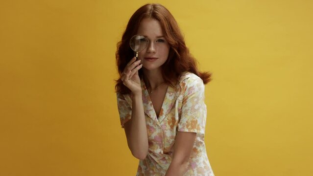 A young white ginger woman on a yellow background looking through a magnifying glass