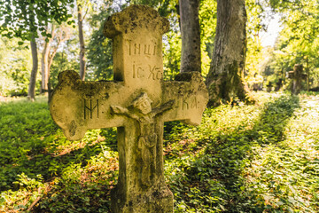 Abandoned greveyard from 1800s. Close up of headstone in the shape of a cross with sculpted jesus figure covered in moss. Sunlit green forest. Horizontal shot. High quality photo