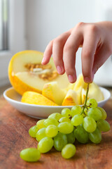 a woman's hand with green grapes, plate of melon slices on a wooden background, concept of fresh fruits and healthy food
