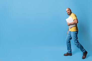 Full length body size view of attractive bearded smile middle-aged man going using laptop over bright blue color background. Casual clothing , yellow t-shirt and blue jeans.