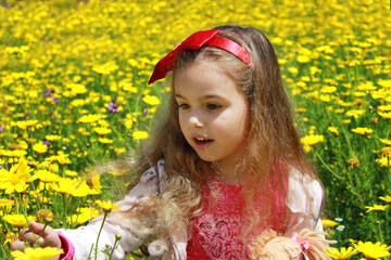 Curly little girl with a red bow in her hair. Girl with a dol on a green meadow among yellow flowers