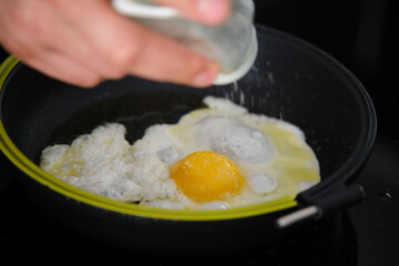 Male hand adding salt to egg in olive oil in frying pan.