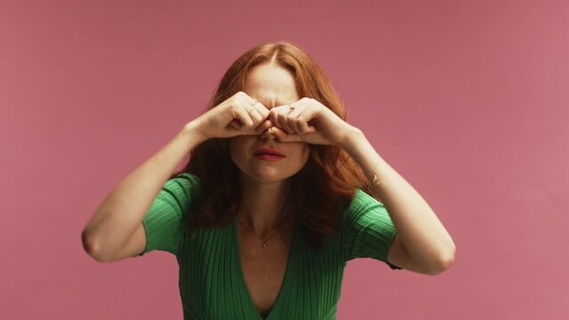 A young white ginger woman, using hand binoculars, rubbing eyes in disbelief and amazement, in front of a pink background wearing a green dress