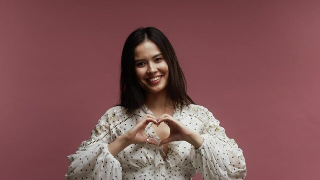 A young asian woman on a pink background forming heart from her hands 