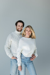 cheerful man standing with hand in pocket and hugging blonde girlfriend in sweater isolated on grey