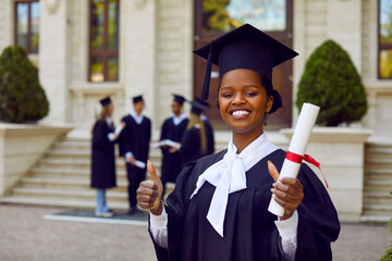 Portrait of joyful female university student showing excellent sign while standing with diploma scroll. Happy african american girl in academic gown and academic cap showing thumbs up outdoors.
