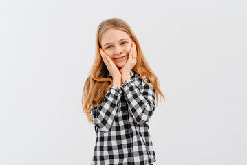 Beautiful young girl with blonde hair smiles at camera, posing, touching face with her hands, wears casual shirt. Teen girl looking happy and delighted. Indoor studio shot on white background