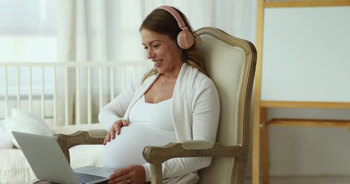 Pregnant woman in headphones sits in armchair using laptop having video call conversation to family, talk to obstetrician-gynecologist, receive professional medical on-line consultation, prenatal care
