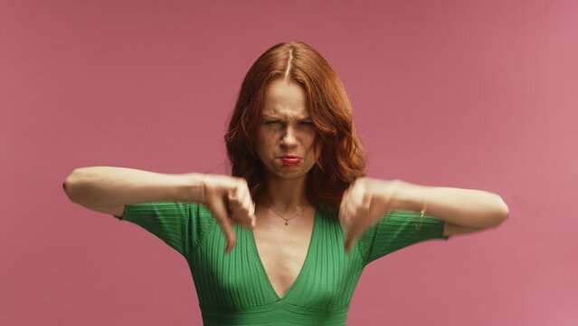 A young white ginger woman, putting thumbs down, disagreeing, in front of a pink background wearing a green dress