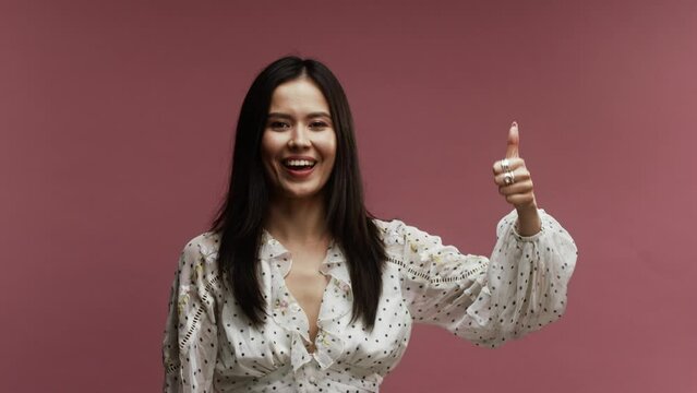 Asian woman wearing a white and spotted dress, raising her hand and giving the thumps up, against a pink background