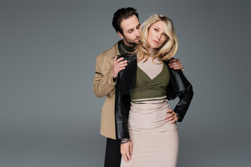 stylish bearded man in sunglasses embracing blonde model in black leather jacket posing isolated on grey
