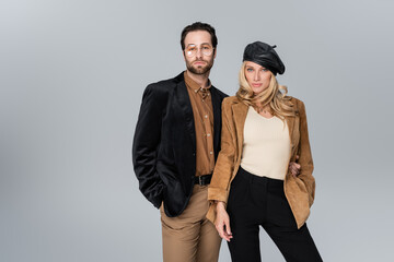 blonde woman in stylish beret posing with bearded man in sunglasses isolated on grey