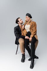 full length of blonde woman in stylish beret sitting on knees of bearded man in sunglasses on grey