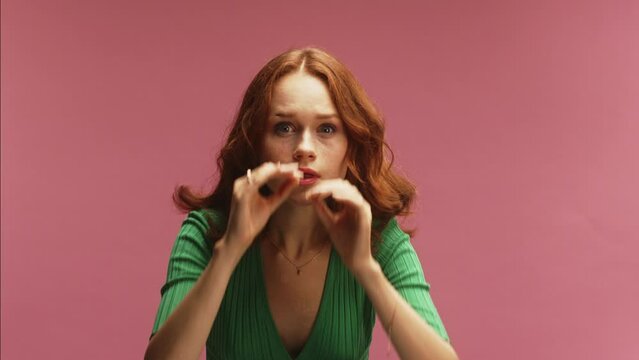 A young white ginger woman, using hand binoculars, rubbing eyes in disbelief and amazement, in front of a pink background wearing a green dress
