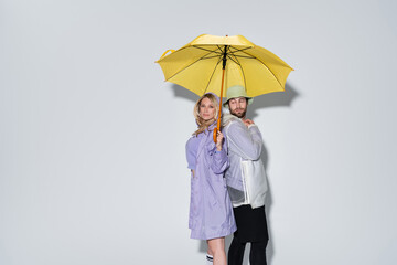 blonde woman in tartan skirt standing with bearded man in panama hat under yellow umbrella on grey