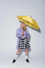 full length of happy woman in tartan skirt and longs socks with sneakers standing under umbrella on grey