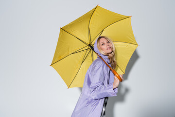 blonde woman purple rain jacket standing under yellow umbrella on grey
