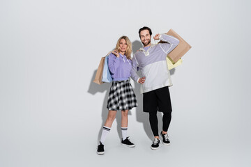full length of joyful man and cheerful woman in tartan skirt holding shopping bags on grey