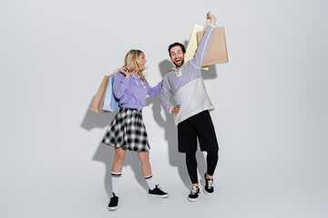 Naklejka premium full length of excited man and cheerful woman in tartan skirt holding shopping bags on grey
