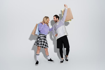 full length of excited man and happy woman in tartan skirt holding shopping bags on grey