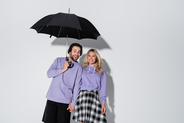 happy couple in purple sweatshirts smiling while standing under umbrella isolated on grey