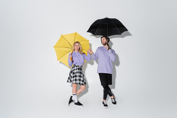 full length of pleased couple in purple sweatshirts holding hands while standing under umbrellas on grey