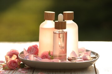 Bottles of rose essential oil and flowers on wooden table outdoors
