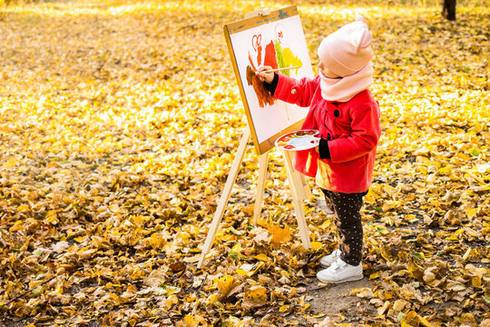 A little girl in a red coat paints a watercolor on the easel in the park against the background of the autumn landscape