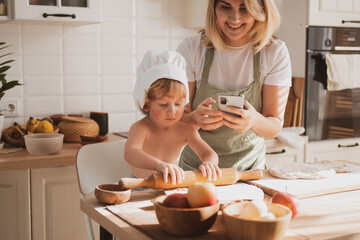 Pretty woman and her little son are preparing pie at home in kitchen and takes pictures on phone