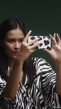 A young Asian woman, wearing a zebra pattern dress, recording a video with her cow print phone, getting a good picture, in front of a green background
