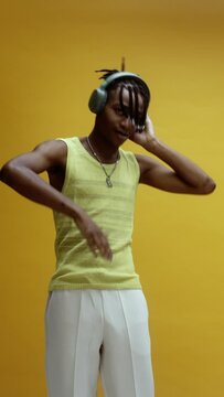 A young stylish black man, wearing a vest and white trousers, enjoying some music on his headphones, in front of a yellow background