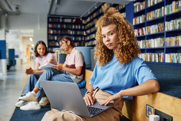 Portrait of curly haired young woman using laptop in college library and looking at camera with...