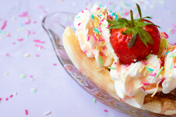 Delicious classic British dessert Ice cream sundae banana split in classic glass bowl with whipped cream, colourful sprinkles, sauce and a strawberry. sprinkles mess close up against white background