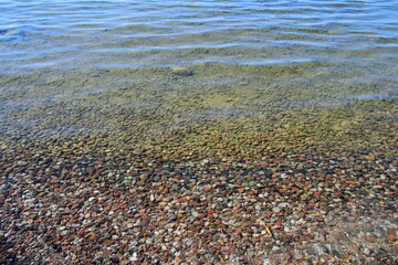 A close up on a vast yet shallow river or lake full of small rocks, stones, pebbles, and other items seen next to the sandy beach on a sunny summer day on a Polish countryside
