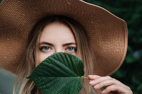 Portrait of young 30 woman caucasian appearance in straw hat with linden leaf closing the mouth. Natural style. Pretty face with big blue eyes. Clean healthy skin. Stylish look. Summer time vacation