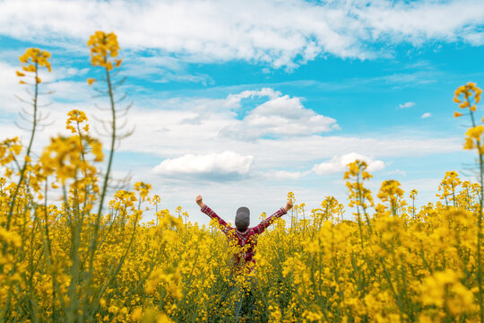 Successful Female Farmer Wearing Plaid Shirt And Trucker's Hat With Arms Raised In The Air In Victorious Pose Standing In Blooming Rapeseed Field And Looking At Horizon