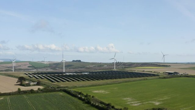 Wind Turbines With Solar Panels In Green Fields Near Saint Newlyn East Countryside In Cornwall, England, United Kingdom. Aerial Wide Shot