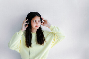 Brunette teen girl wearing headset standing against white cement wall and listening music. Lifestyle and leisure.
