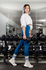 Fototapeta na wymiar Sporty fitness girl is posing at the dumbbells in the gym