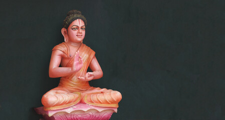 The Buddha sits in a lotus position on a black background with his hands clasped in the Vitarka...