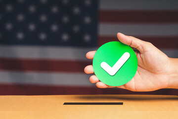 Hand holding a green circle paper with white check mark symbol overhead the voting box with an American flag background