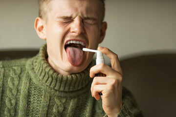 Sick caucasian young man with open mouth and long tongue using a throat spray against a dry cough....